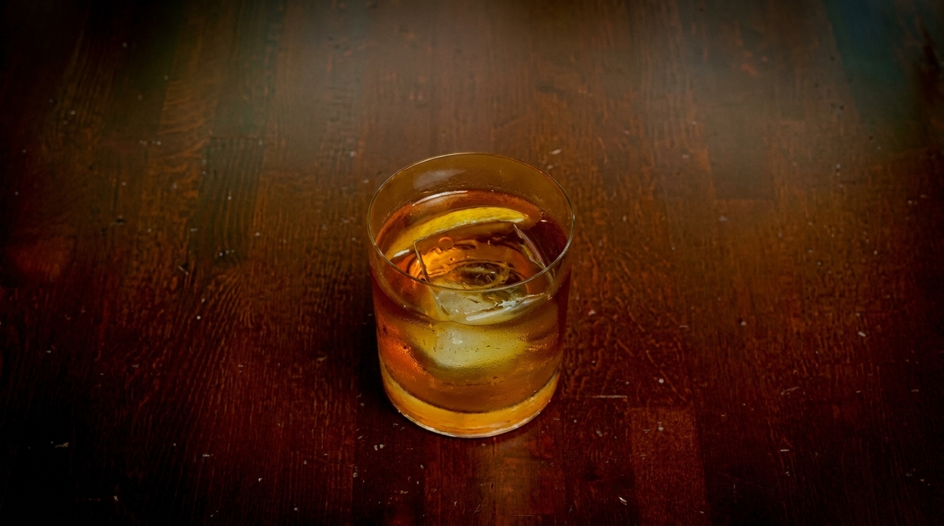An amber cocktail with a large ice cube and citrus peel sits on a dark wooden table.