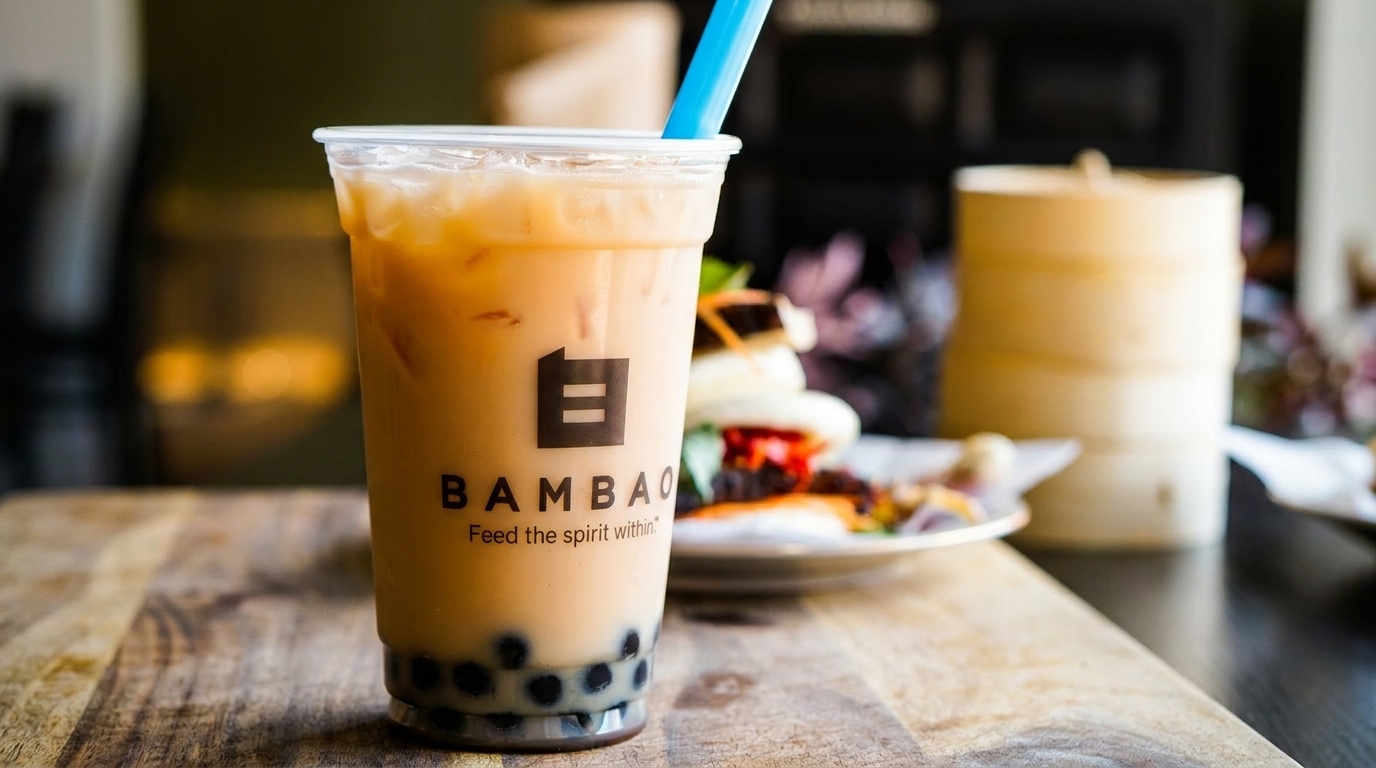 A BambaO bubble tea with a blue straw, bao buns, and a bamboo steamer on a wooden table.