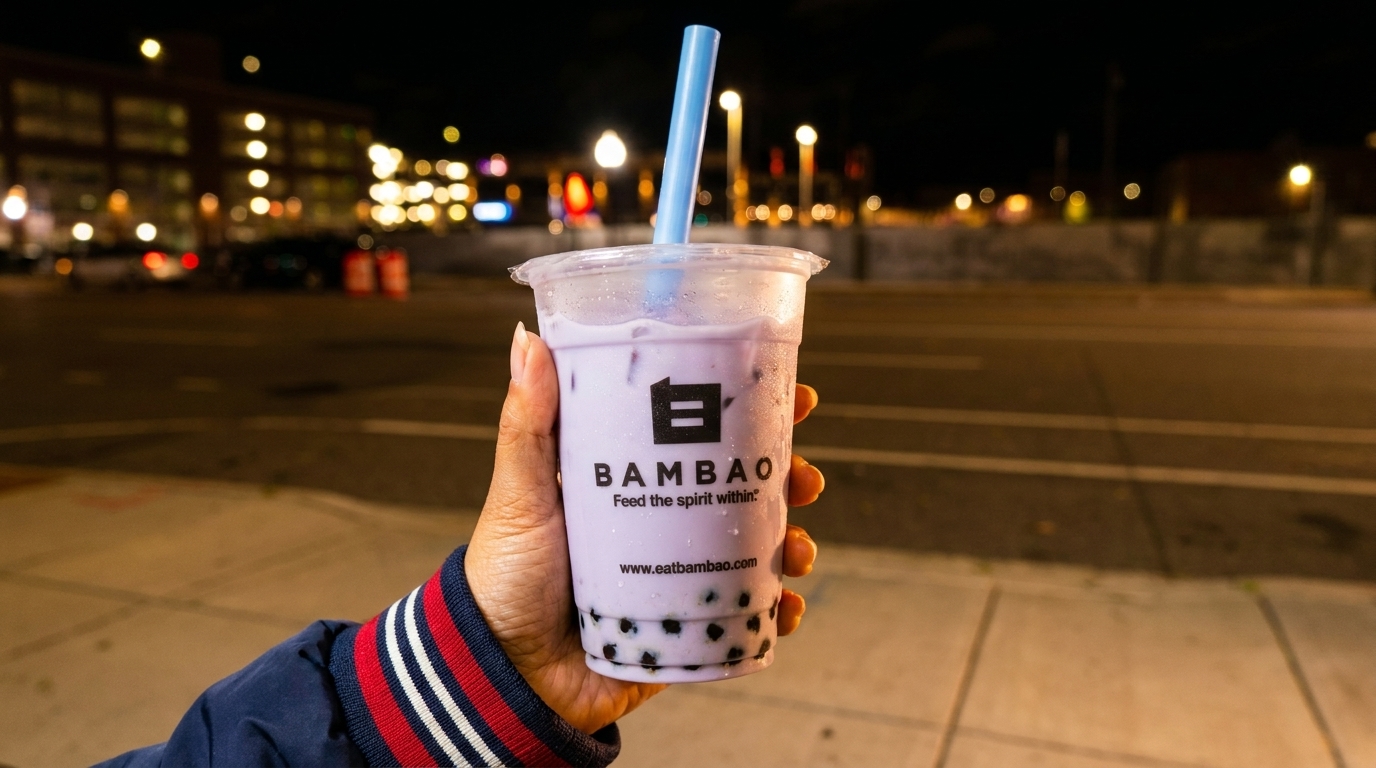 Hand holding a Bambao taro bubble tea with a blue straw against a blurred city night background.