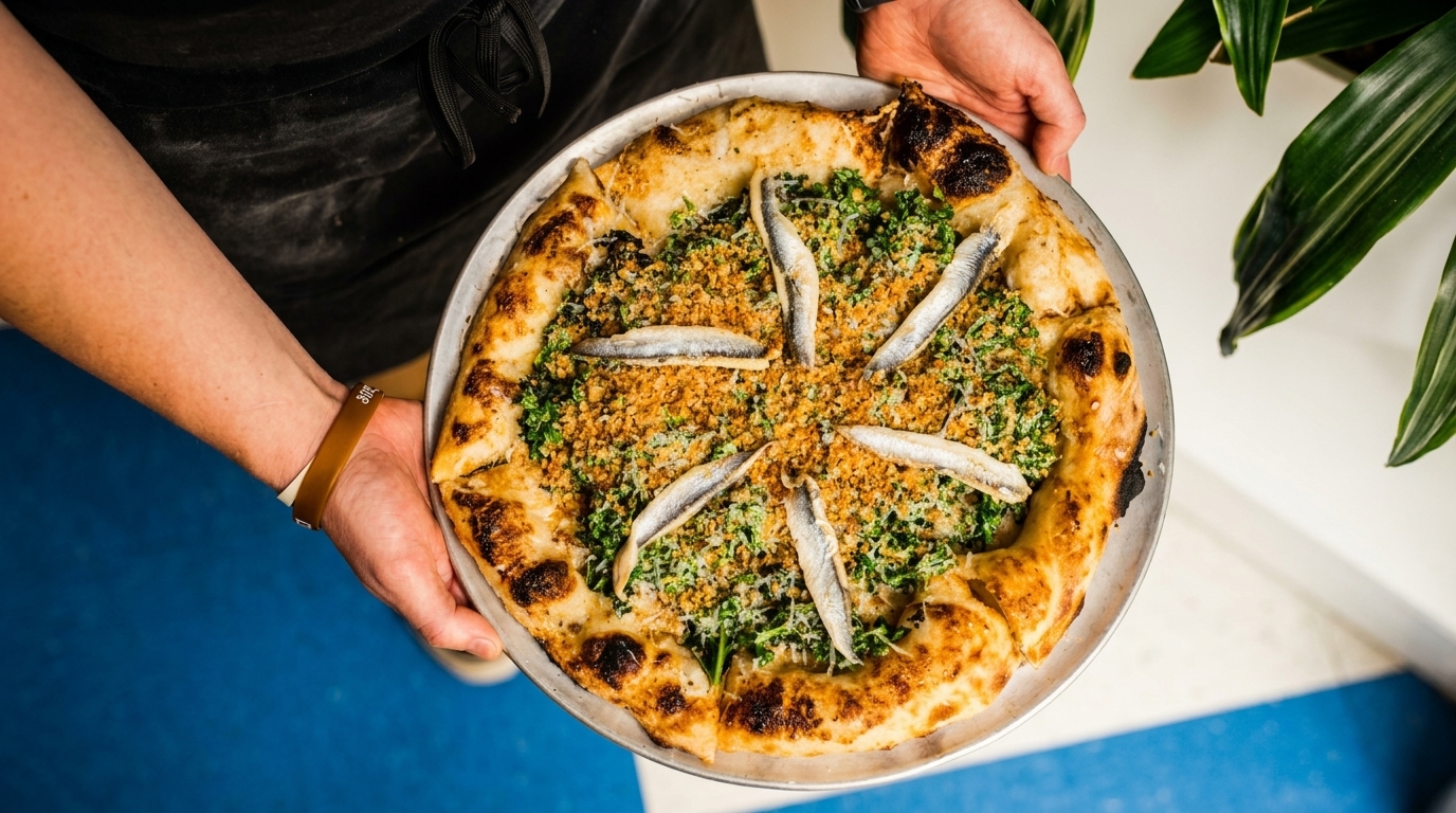 Hands holding a pizza topped with anchovies, breadcrumbs, and greens on a metal tray.