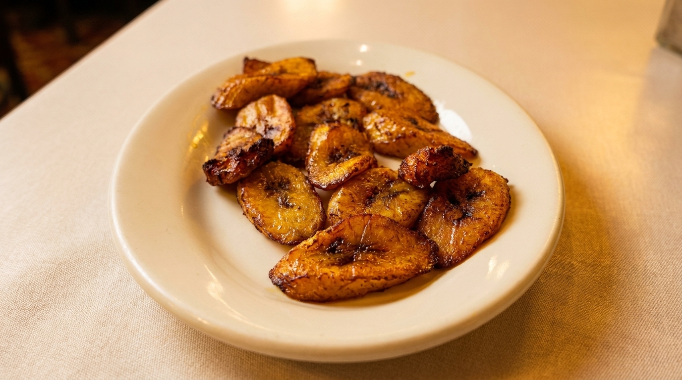 A white plate filled with golden-brown fried plantains, some with caramelized edges, on a light tablecloth.