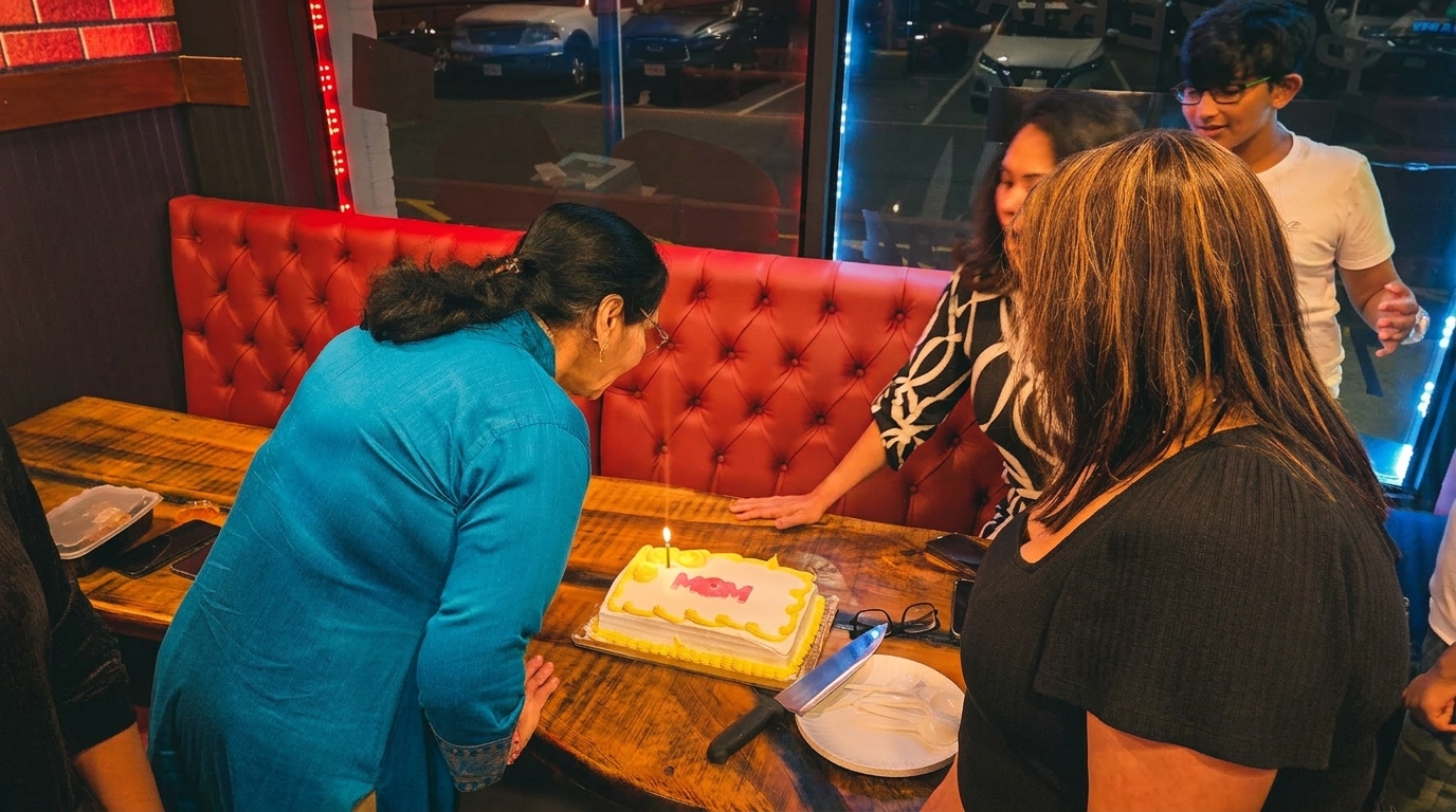 A woman in blue blows out a candle on a "Mom" birthday cake at a restaurant booth.