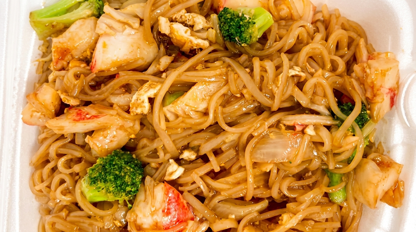 Close-up of crab pad see ew with flat noodles, broccoli, and onions in a white takeout container.