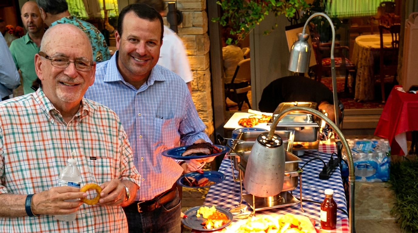 Two smiling men at a buffet table with barbecue ribs, onion rings, and other food.