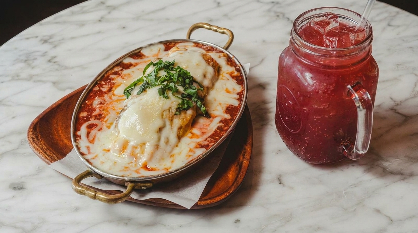 Baked pasta dish with melted cheese and basil, next to a red iced drink on a marble table.