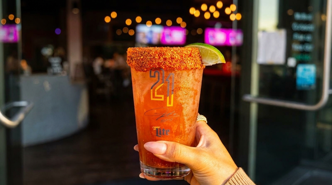 Hand holding a michelada with a chili-salted rim and lime wedge, in front of a restaurant entrance.