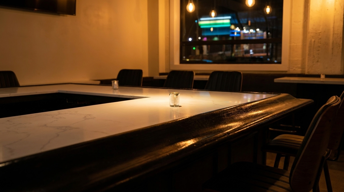A dimly lit bar area with a white marble countertop, dark chairs, and a street view outside.
