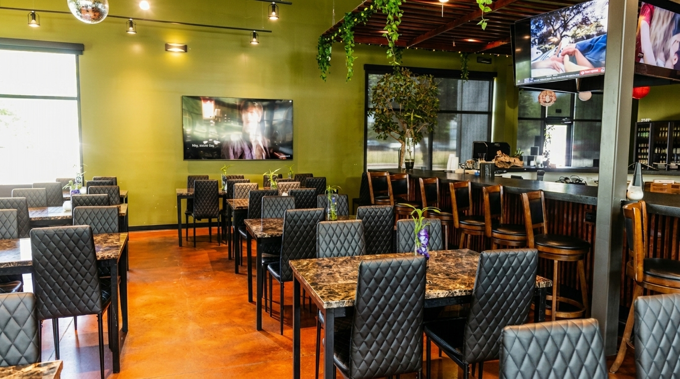 Restaurant interior with rows of black chairs at marble-top tables, a bar with stools, and TVs on olive green walls.
