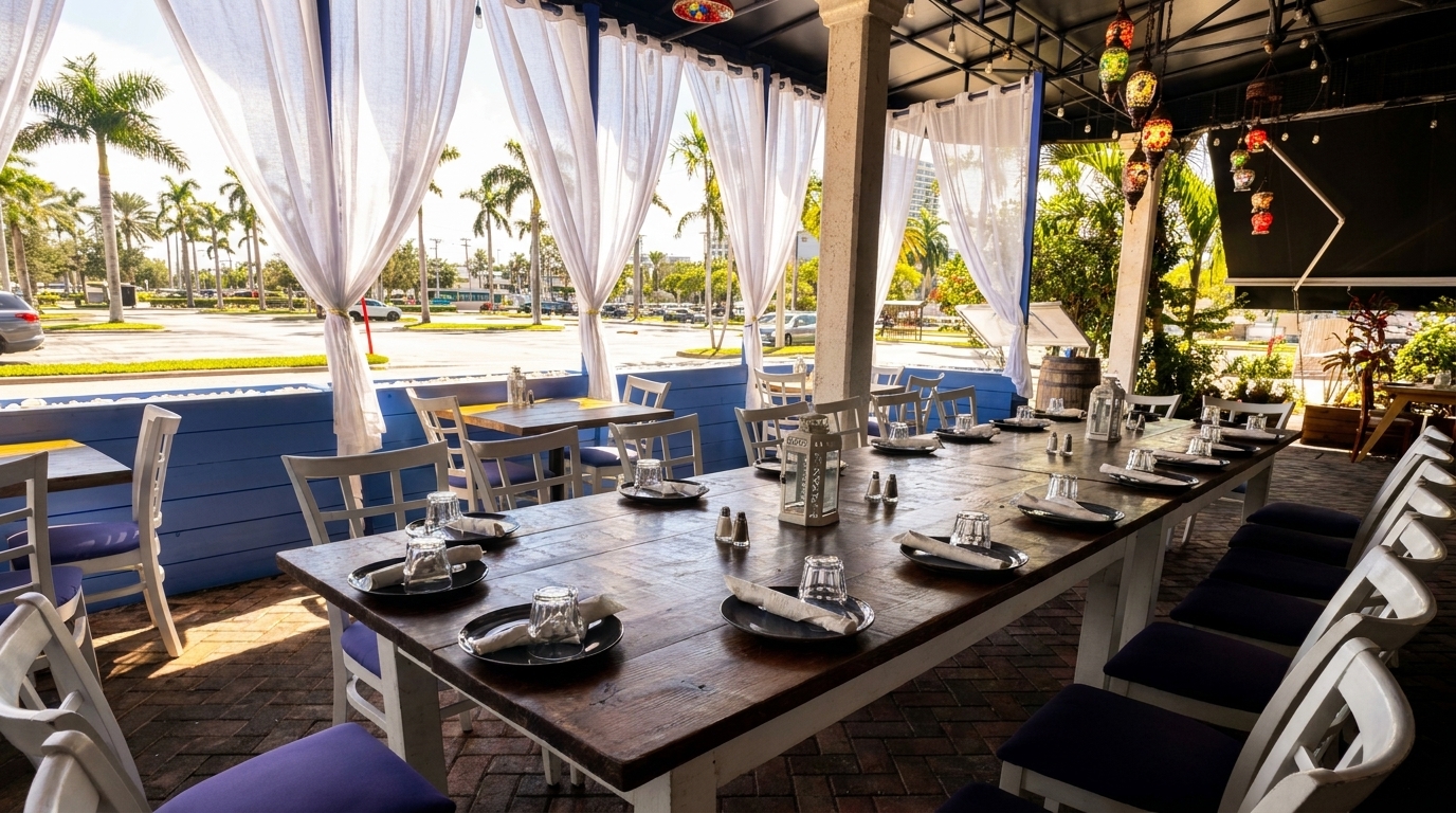 Outdoor restaurant patio with a long wooden table set for dining, white chairs, and palm trees in the background.