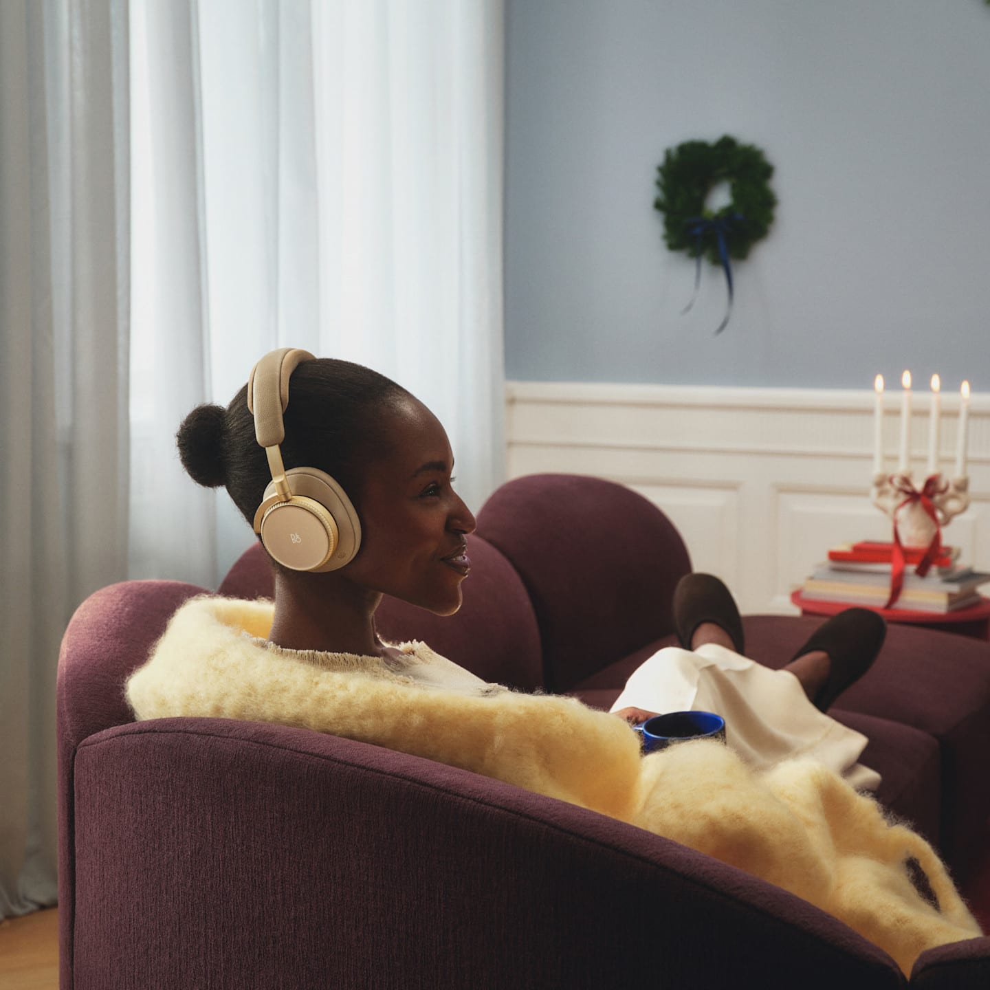 Person wearing gold Super Hoodie apparel relaxing on purple couch with blanket in cozy room with candles and holiday decor.