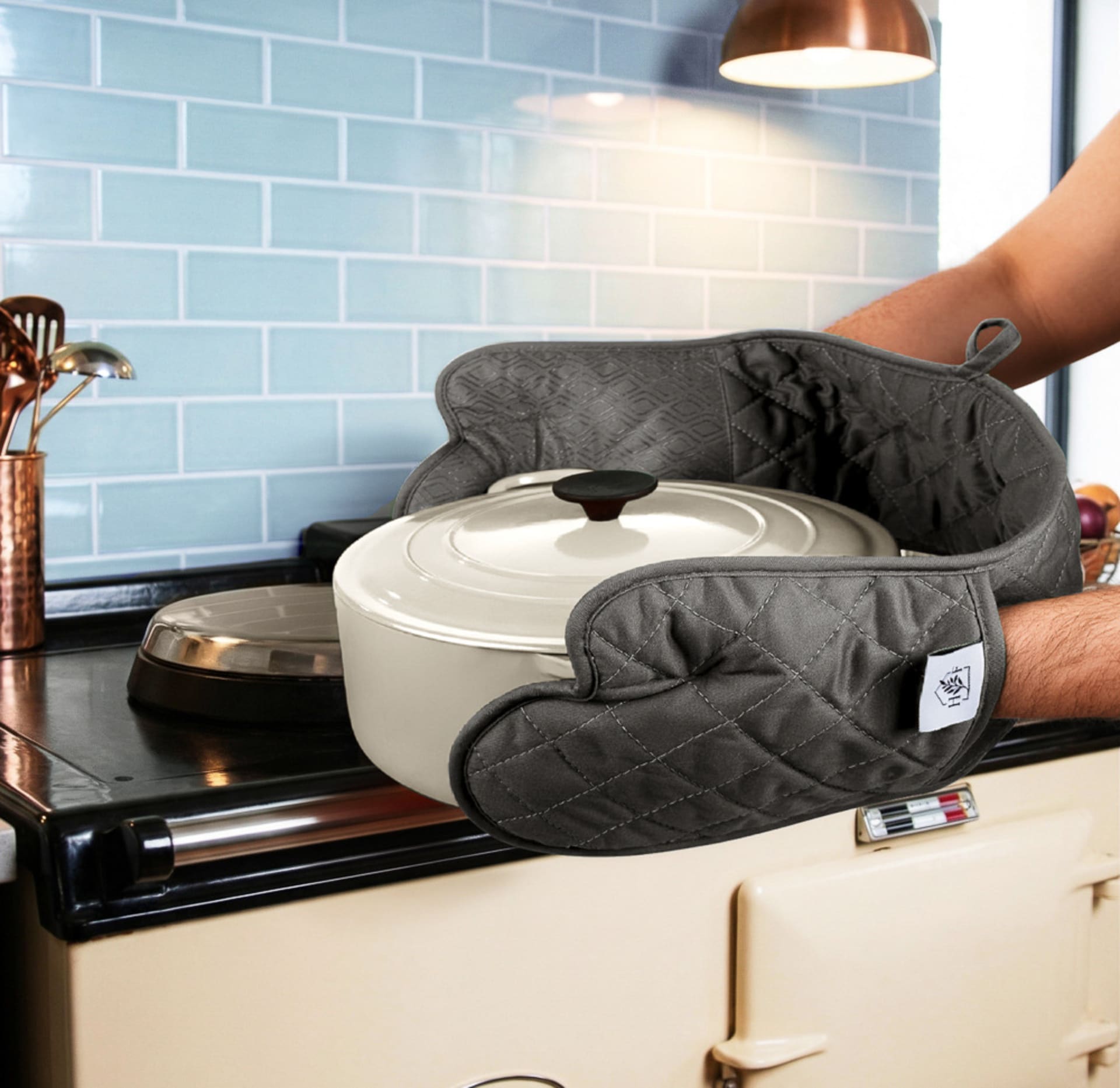 Grey Homefolk oven gloves holding a white casserole pot beside a cream range cooker and pale blue tiled wall