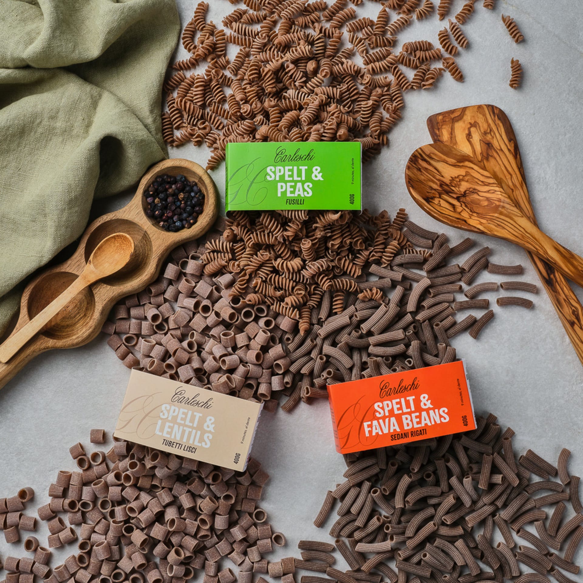 Three varieties of Carleschi pasta arranged with their labels, wooden utensils and green linen on a grey surface