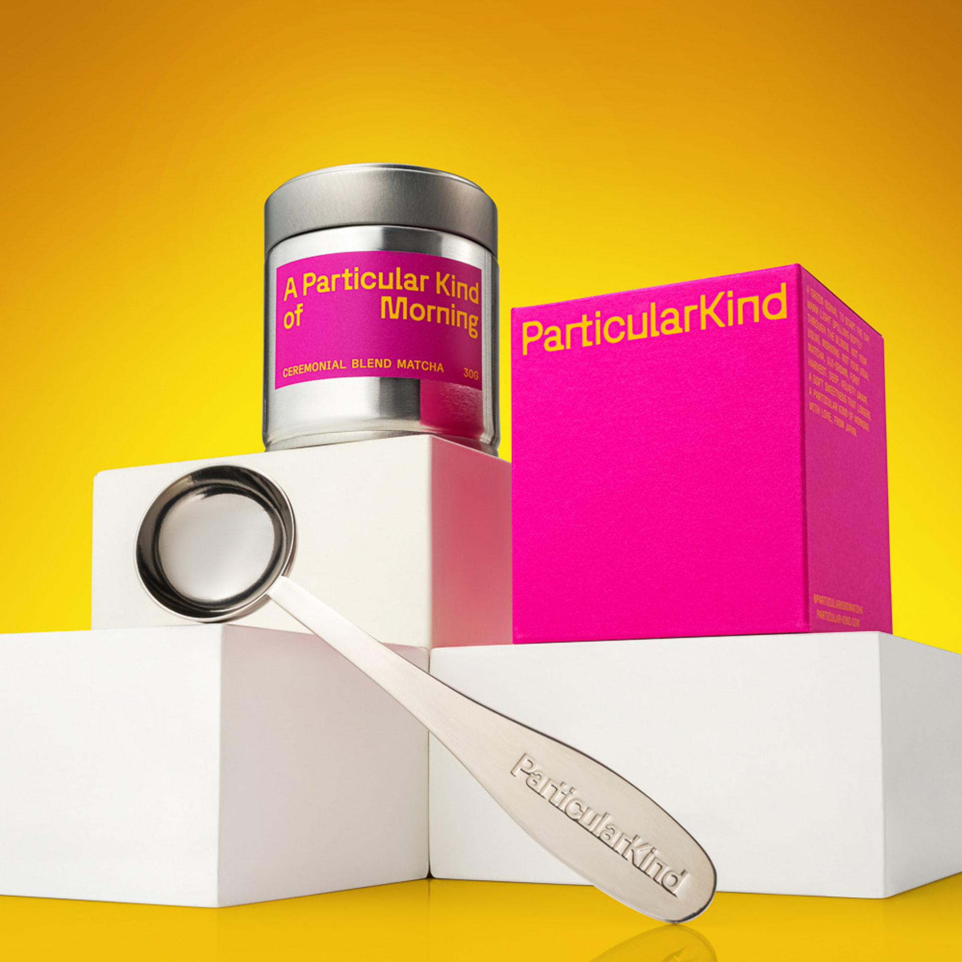 Particular Kind matcha tin, pink box and silver scoop arranged on white plinths against a yellow background
