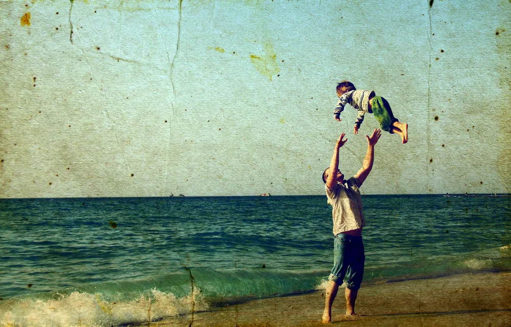 A man on a beach throws a child into the air as waves roll in, with a textured, vintage filter across the scene.