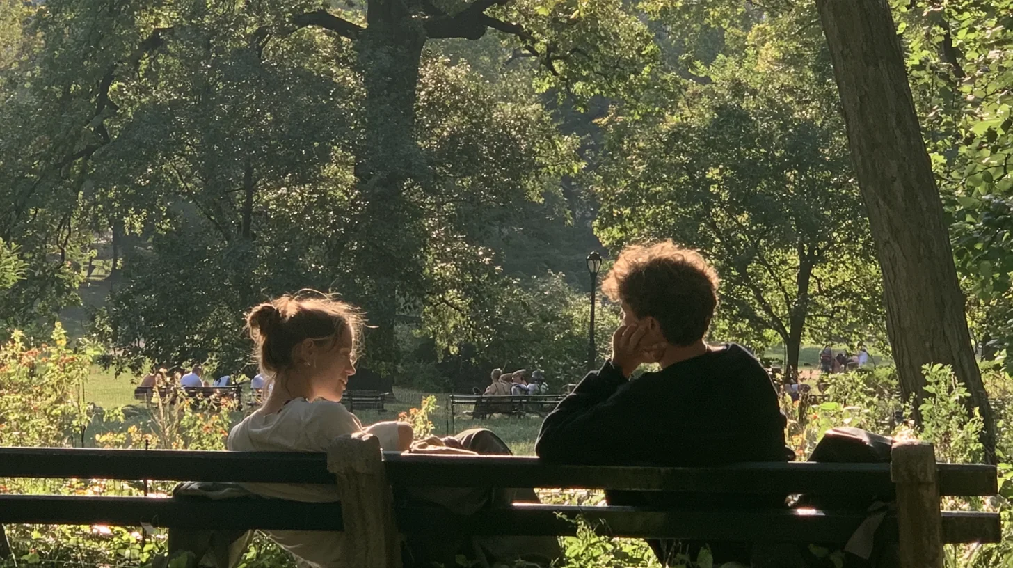 Two young people sit side by side on a park bench in warm afternoon light, the woman smiling as she talks and the man resting his chin on his hand, listening, with trees behind them.