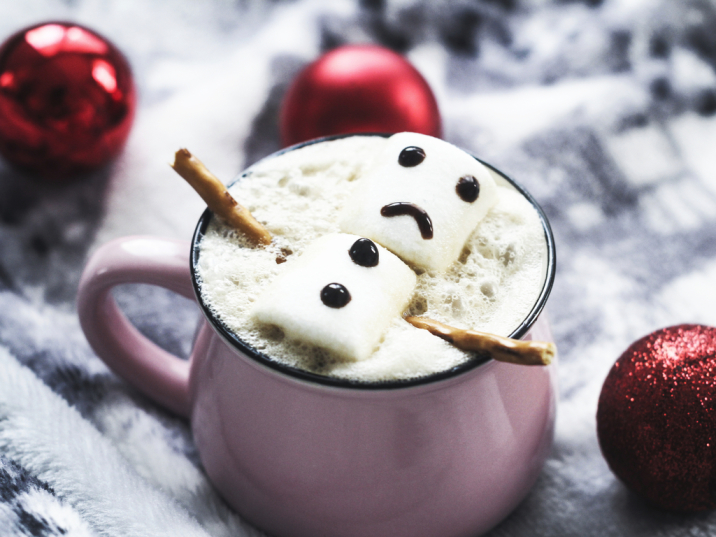 Marshmallow figure with a sad face floats in hot chocolate, decorated with pretzel sticks, surrounded by red ornaments.