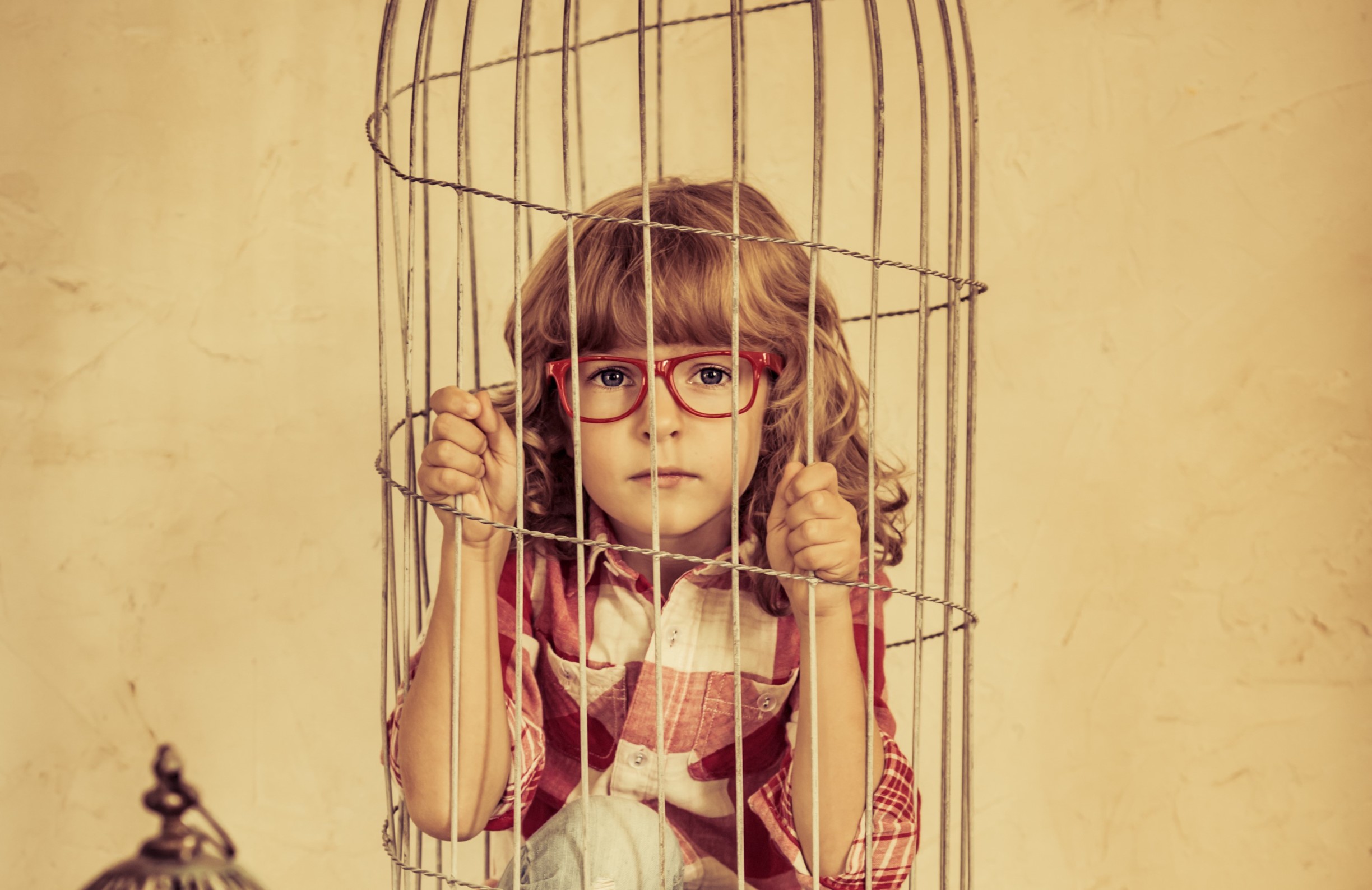 Young child with curly hair and glasses, sitting in a birdcage, symbolizing feelings of restriction during toddler development.
