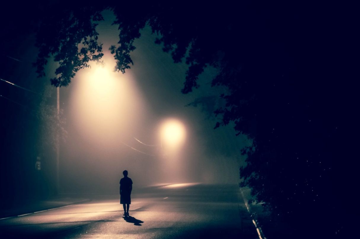 Figure standing alone on a foggy street illuminated by streetlights, reflecting themes of solitude and resilience.