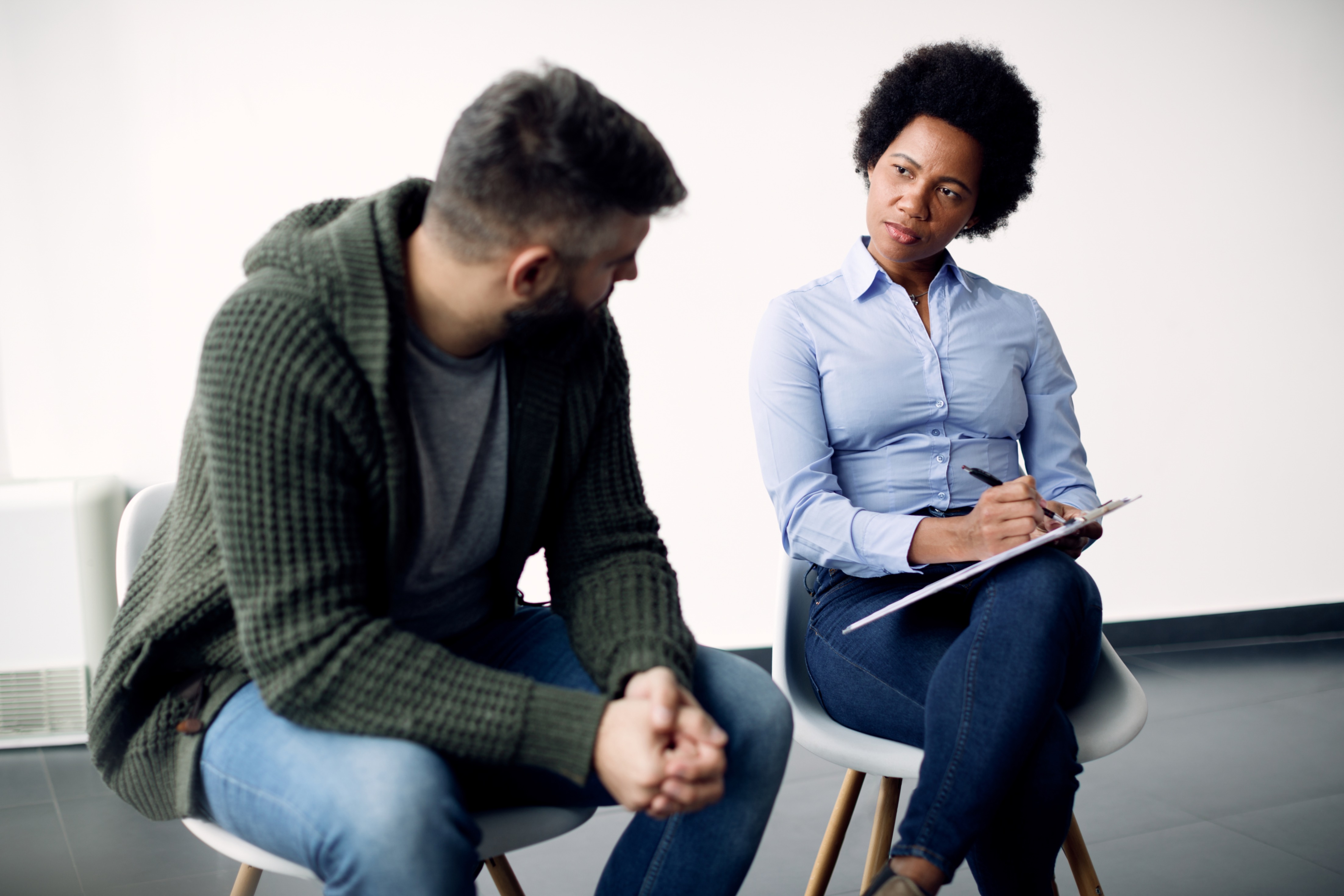 A female therapist with a clipboard listens to a patient speaking during a therapy session.