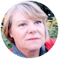 A woman with short hair, wearing a red sweater, looks thoughtfully among vibrant flowers, reflecting a supportive therapy approach.