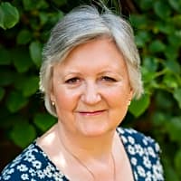 Smiling woman with short gray hair, wearing a flower-patterned top, surrounded by green foliage, representing a supportive therapist.