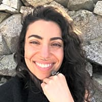 Smiling woman with curly hair seated against a stone wall, reflecting a creative and relaxed spirit.