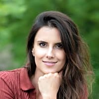 Young woman with long brown hair resting her chin on her hand, smiling against a green outdoor background.