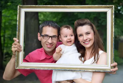 Happy family portrait with parents holding their smiling baby in a picture frame surrounded by greenery.