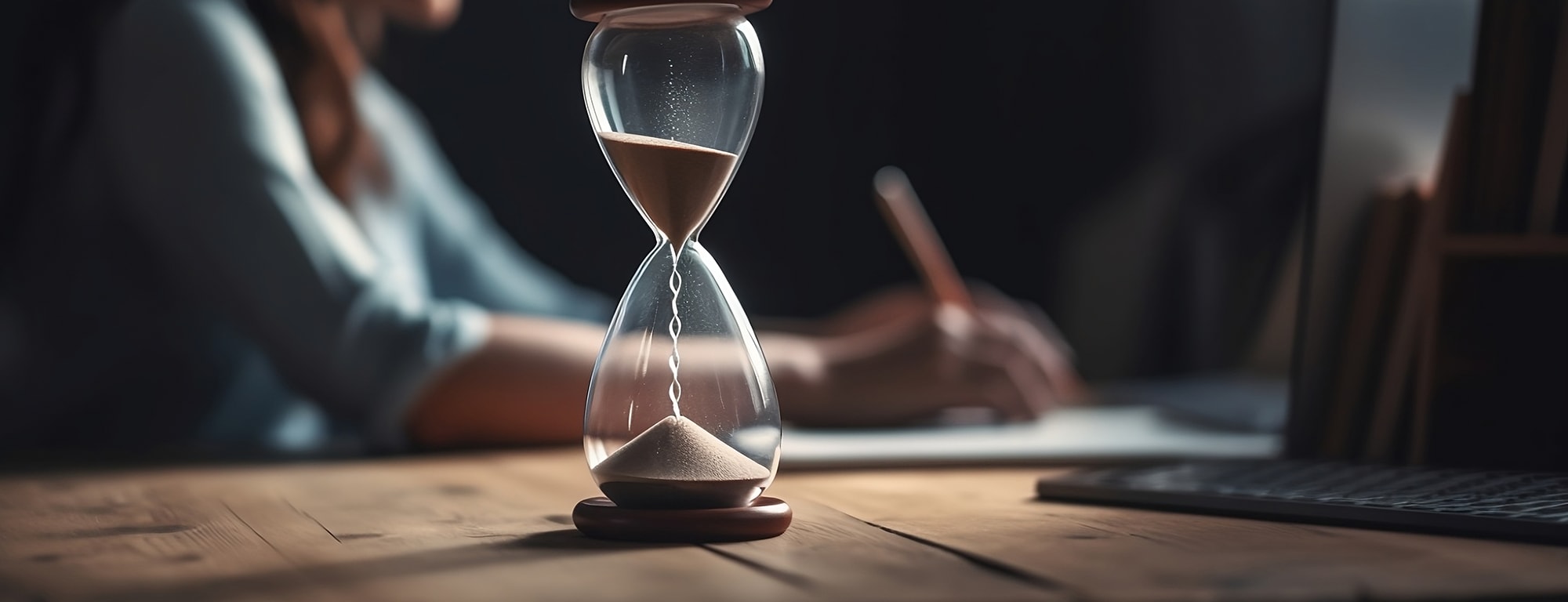 Close up of hourglass with sand flowing down. Blurred woman at laptop in background.
