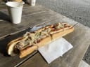 Grilled bratwurst with sauerkraut and two sauces in a toasted bun on a rustic wooden table, representing a butcher's offering at Farmers' Market Taranaki.