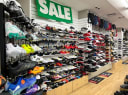 Rows of athletic shoes, cleats, and casual footwear displayed on shelves in a retail shoe store with a prominent 'SALE' sign.