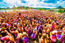 Vibrant crowd covered in colourful powder celebrating outdoors under a blue sky, possibly a Holi festival.