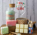 Assortment of artisan crafts: layered bath salts, pink handmade soap, a wine glass, gold candle, wax melts, and car diffuser.