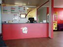 Interior of Ngatea Pizza shop featuring a large menu board, red service counter with a pizza chef logo, and arcade games.
