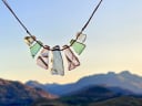 Handcrafted sea glass necklace featuring unique green and clear pieces, suspended against a vibrant sunset sky and blurred Queenstown mountains. Showcases artisan jewellery.