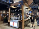 Charing Cross Cheesery stall with 'All roads lead to cheese' sign at bustling Riverside Market, with a gelato stand in the foreground.