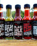 Five bottles of The Chilli Co's artisan hot sauces, including Black Garlic Sriracha and Original Hot Pepper Sauce, on a wooden table.