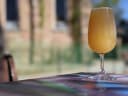 A tall glass of hazy golden beverage sits on a vibrant, multi-coloured table outdoors, with a blurred building in the background.