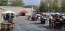 Busy outdoor patio scene with people dining at tables, a white tent, a red DJ tent, and cars in the background.