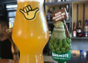 A tall glass of hazy craft beer with a Shaka hand logo, alongside a Behemoth Brewing Company tap handle, in a bar.