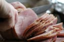 Hands expertly slicing a large, succulent cooked ham with a knife, creating thin, even slices for Waitaki Bacon and Ham.