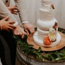 Two people cutting a tiered wedding 'cake' made of Whitestone artisan cheese wheels, decorated with fresh flowers, on a rustic barrel display.