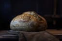 Freshly baked golden sourdough loaf with a scored crust, resting on a dark oven mitt on a wooden surface, from Breads & More at Otago Farmers Market.