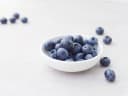 Fresh, plump blueberries in a white bowl and scattered on a light textured surface, showcasing Blue Willow Blueberries' produce.