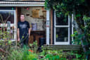 Devils Cups Coffee maker stands in his rustic workshop, with a 'Devils Cups' sign on the brick wall, surrounded by lush New Zealand greenery.