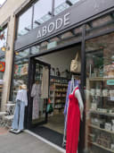 Abode shopfront displaying women's fashion, homeware, and gifts. Mannequins wear dresses and casual outfits. Products are visible through large windows.