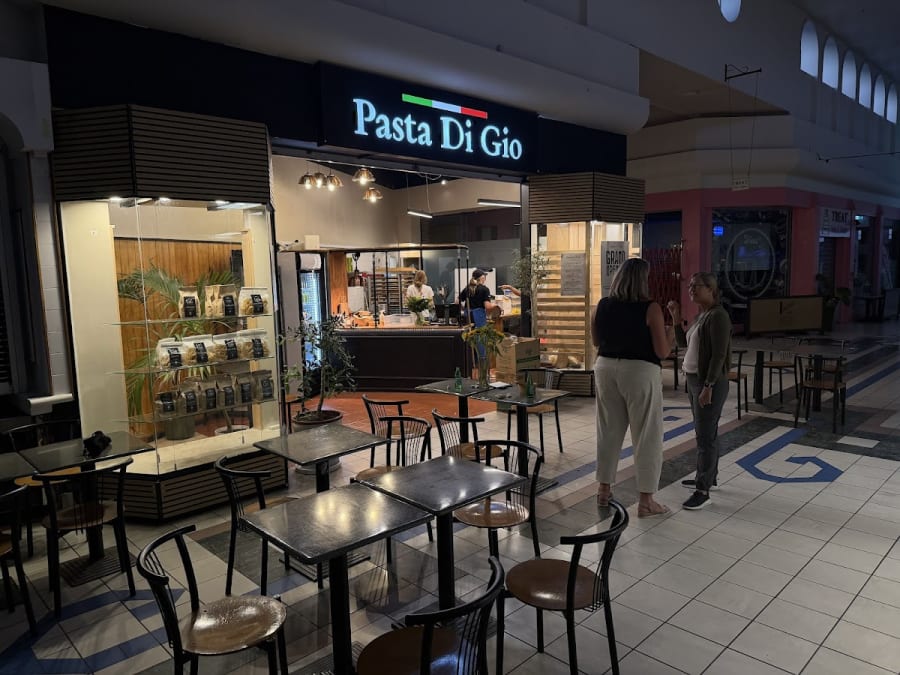 Pasta Di Gio shopfront with Italian flag colours, displaying packaged artisan pasta and staff preparing food. Indoor market setting.