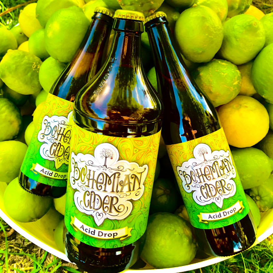 Bohemian Cider 'Acid Drop' bottles with yellow caps nestled among fresh green limes and lemons, showcasing artisan beverages.