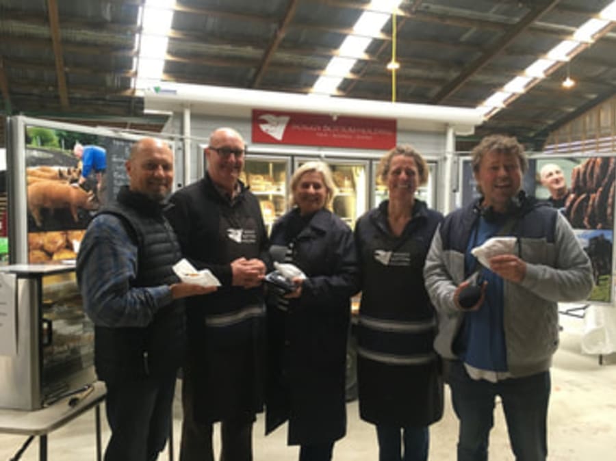 Five people, including Soggy Bottom Holdings butchers, holding meat samples at their stall, with display boards of pigs and products, likely at Cambridge Farmers' Market.