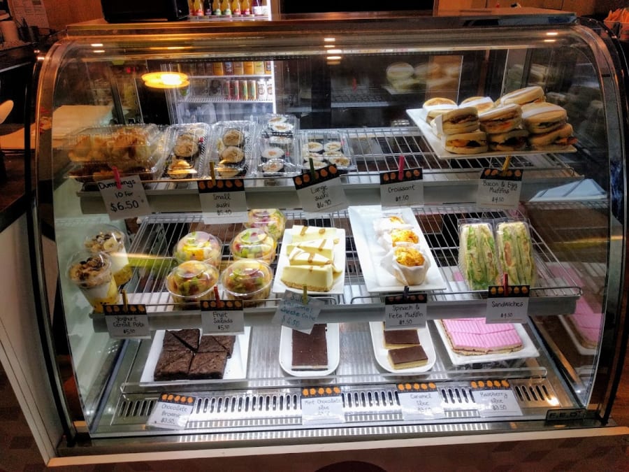 Refrigerated display case filled with various baked goods like croissants, muffins, brownies, and slices, alongside sushi, dumplings, and fruit salads.